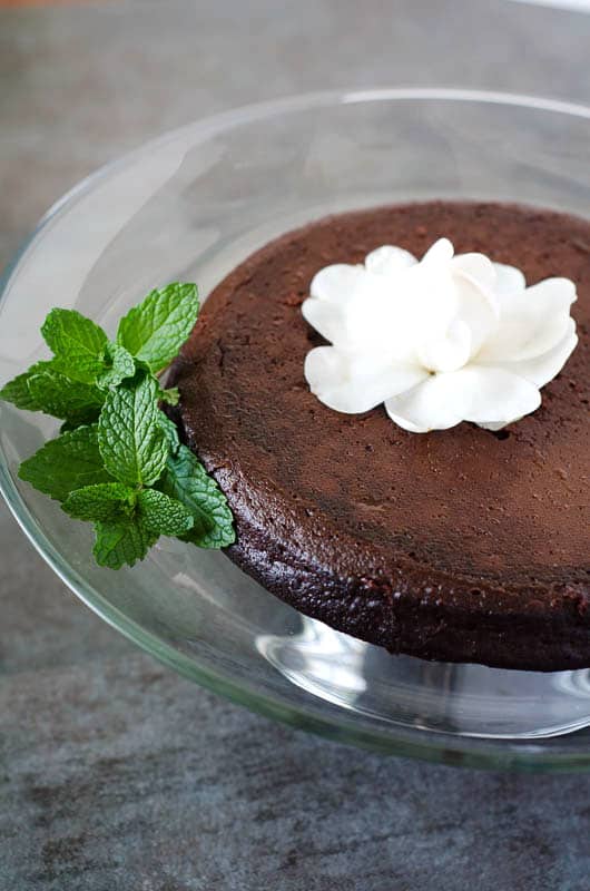 paleo flourless chocolate cake on a clear cake stand topped with a white flower and surrounded by mint leaves