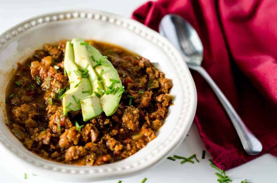 white bowl full of chili topped with avocado slices with red napkin and spoon next to it