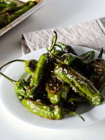 White plate of roasted shishito peppers with pan in the background