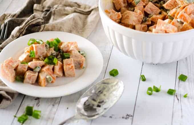 sweet potato salad with bacon and green onions on a white plate with a large serving bowl in background