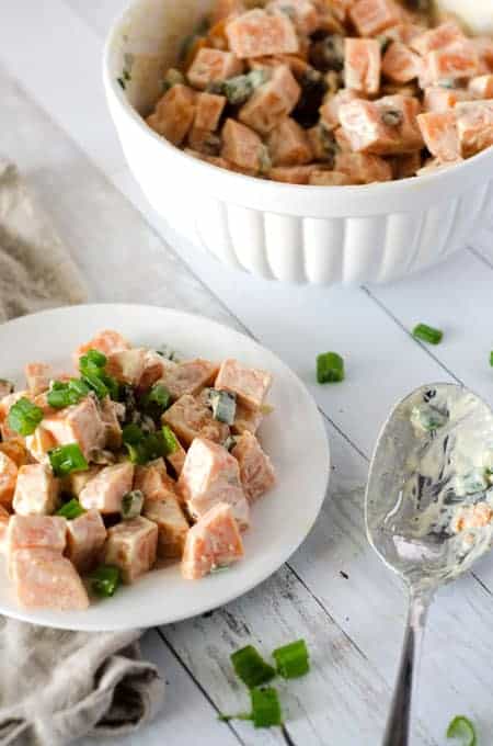 vertical image of sweet potato salad with bacon and green onions on a white plate with a bowl of sweet potato salad in the background