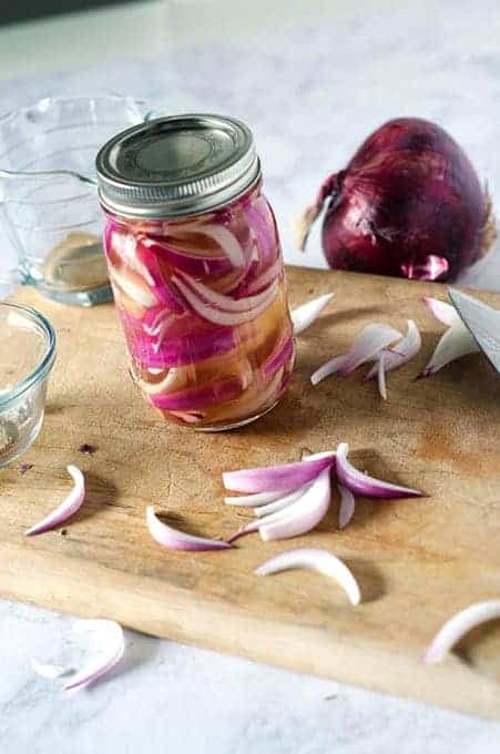 Side view of a jar of pickled red onions on a cutting board with kitchen tools surrounding it