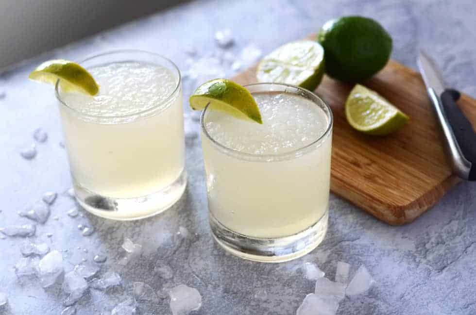 two glasses of frozen limeade surrounded by ice and a cutting board with limes