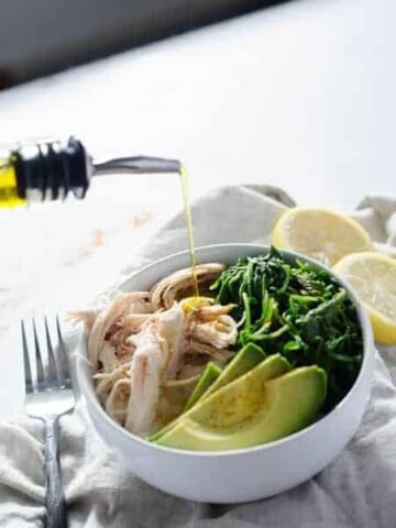 bowl of chicken, greens, and avocado being drizzled with olive oil