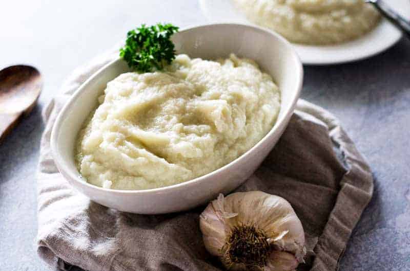 bowl of cauliflower mashed potatoes surrounded by a napkin, head of garlic, and serving spoon