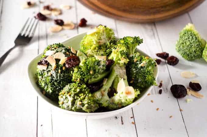 side view of white bowl of raw broccoli salad surrounded by cranberries and almonds with a fork in the background