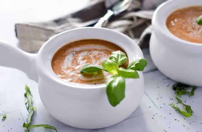 white crock filled with tomato soup with a cream swirl on top and a sprig of basil
