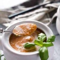 white crock of tomato basil soup with a hand holding a spoonful of soup above the dish