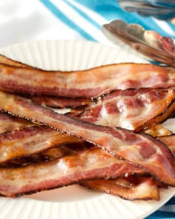 pile of bacon on a white plate on a white and blue striped towel with tongs in the background