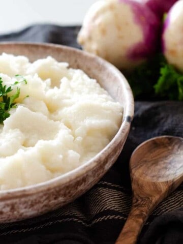 bowl of mashed turnips topped with parsley with a spoon and whole turnips in the background