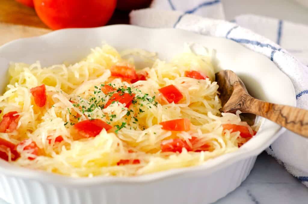 side view of round white dish with cooked spaghetti squash with tomatoes and a wooden serving spoon