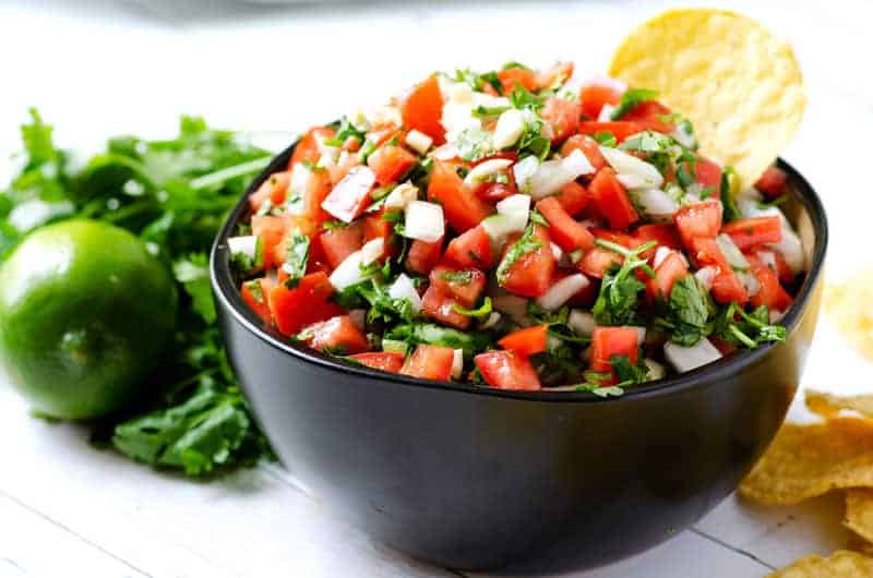 Horizontal image of a black bowl of the best pico de gallo recipe surrounded by tortilla chips
