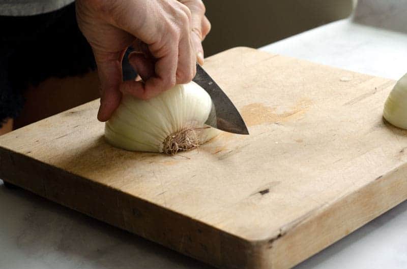 Wooden cutting board with half an onion face down and a woman's hands holding the onion and slicing it with a knife