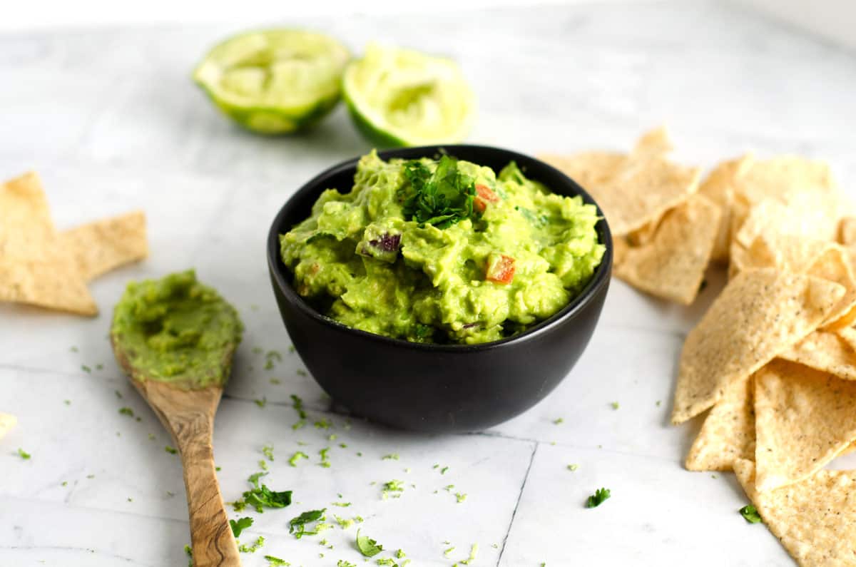 small black bowl of guacamole surrounded by squeezed lime halves, tortilla chips, and a wooden spoon