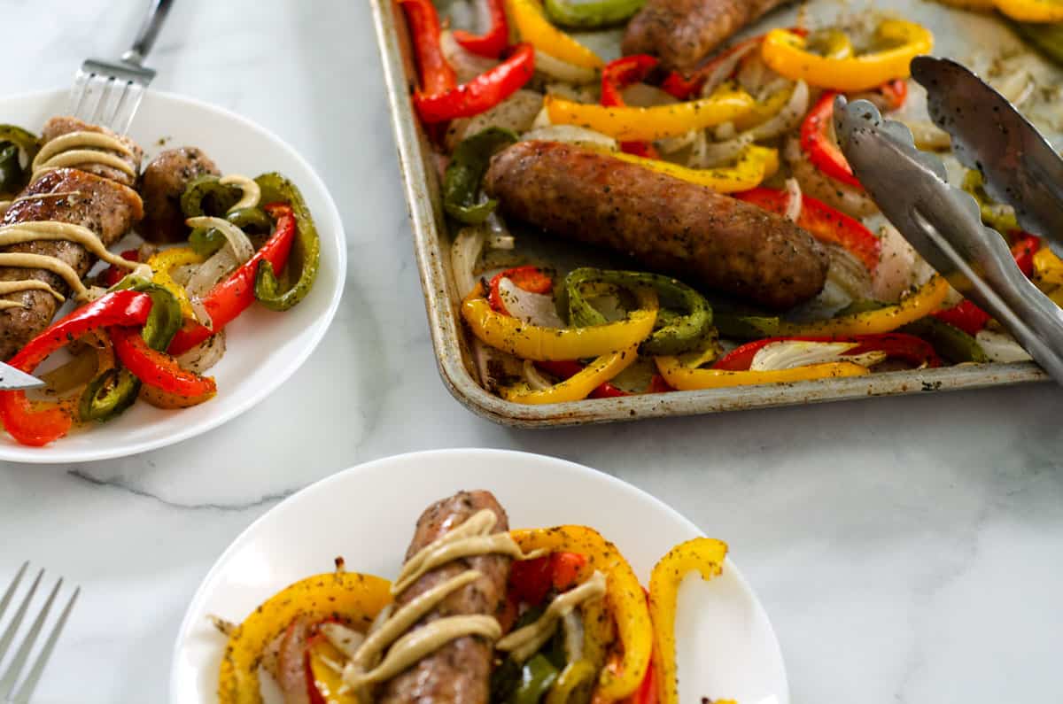 Roasted sausage peppers and onions on a plate drizzled with dijon mustard with a sheet pan in the background