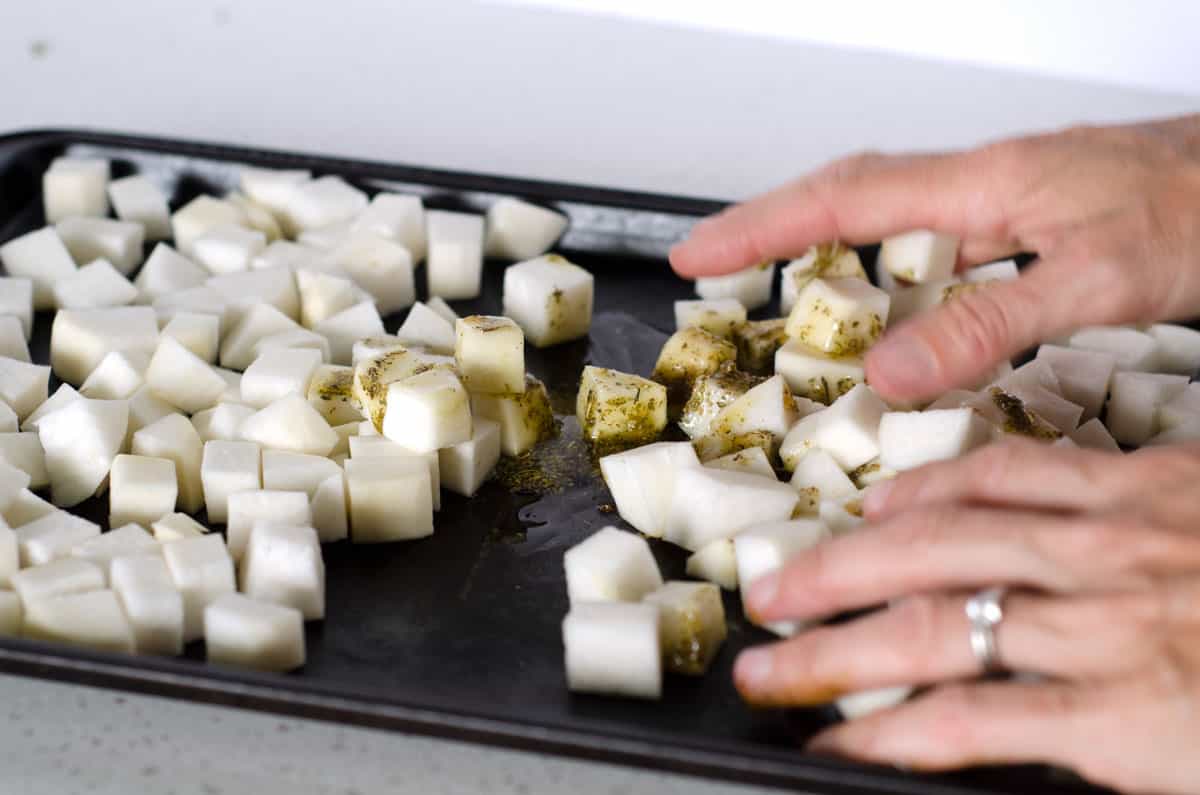 Baking sheet of raw turnip cubes with oil and a pair of hands tossing them together
